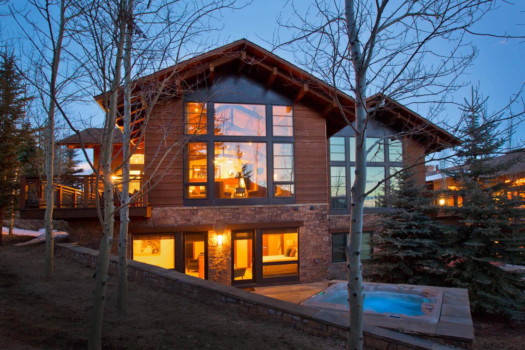 The exterior of a Granite Ridge Residence at dusk with large lit up windows and a hot tub
